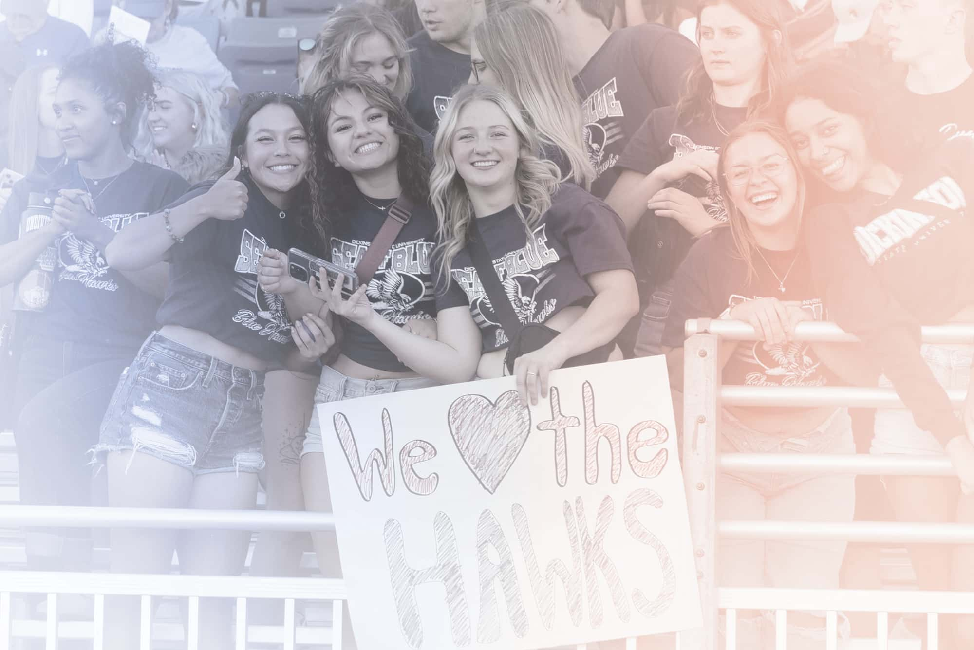 A crowd of students at a game with one student holding a sign that says "We love the Hawks" with a heart instead of the word "love"