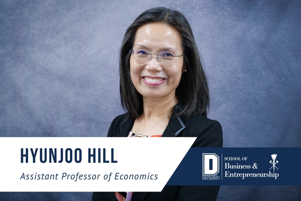 A professional headshot of Hyunjoo Hill, assistant professor of economics at Dickinson State University. She is smiling and wearing glasses and a black blazer over a patterned blouse, standing in front of a blue studio background. A white and navy graphic banner across the bottom of the image displays her name, "Hyunjoo Hill," and her title, "Assistant Professor of Economics," alongside the Dickinson State University School of Business & Entrepreneurship logo.